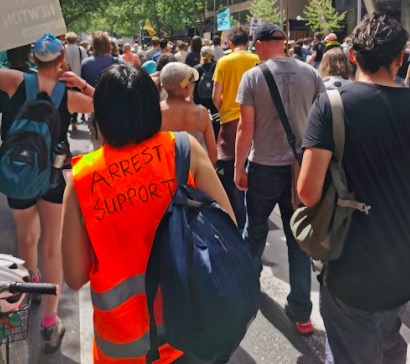Person walking away from camera in a street protest wearing a fluorescent orange vest with the words "Arrest Support" written on it