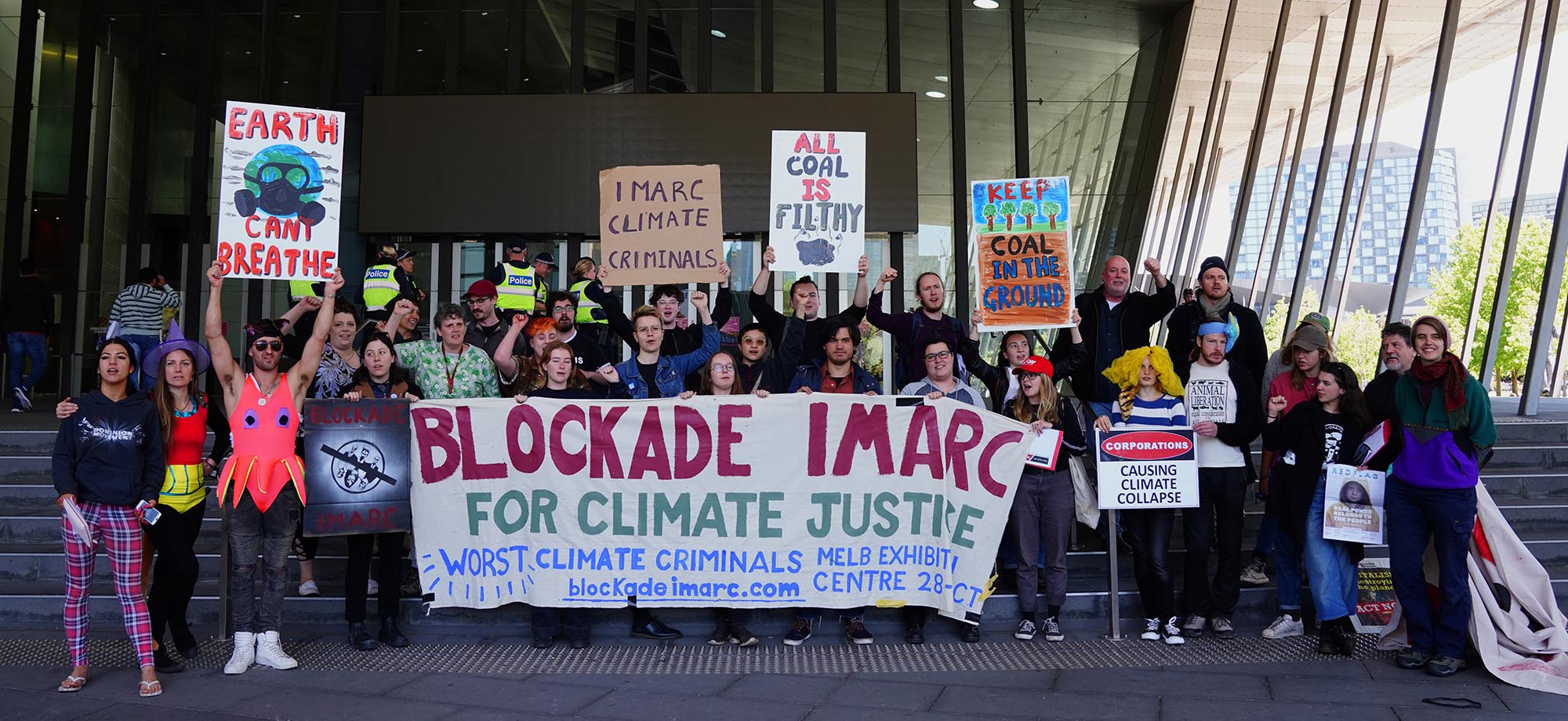 Group of Protestors standing in front of the Exhibition Centre behind a large banner that reads "Blockade IMARC"